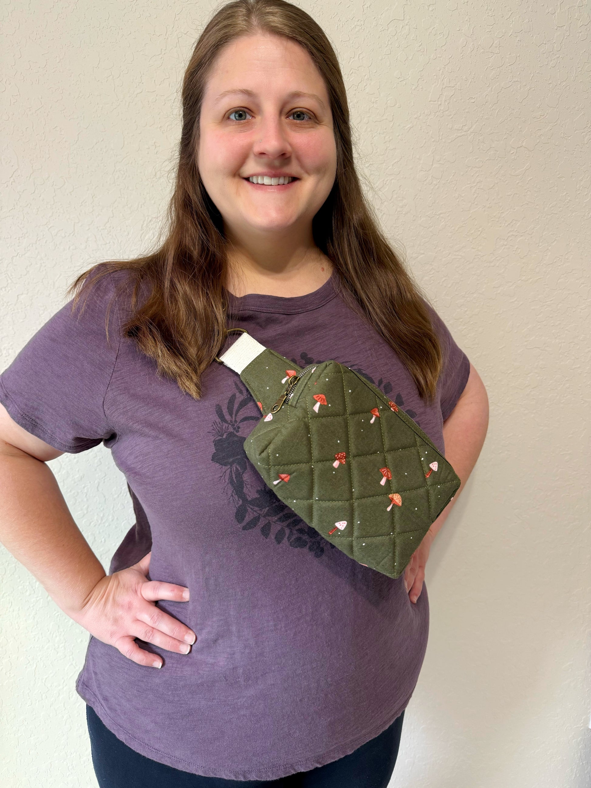 Person wearing a purple shirt with a green quilted bag over their shoulder against a plain background